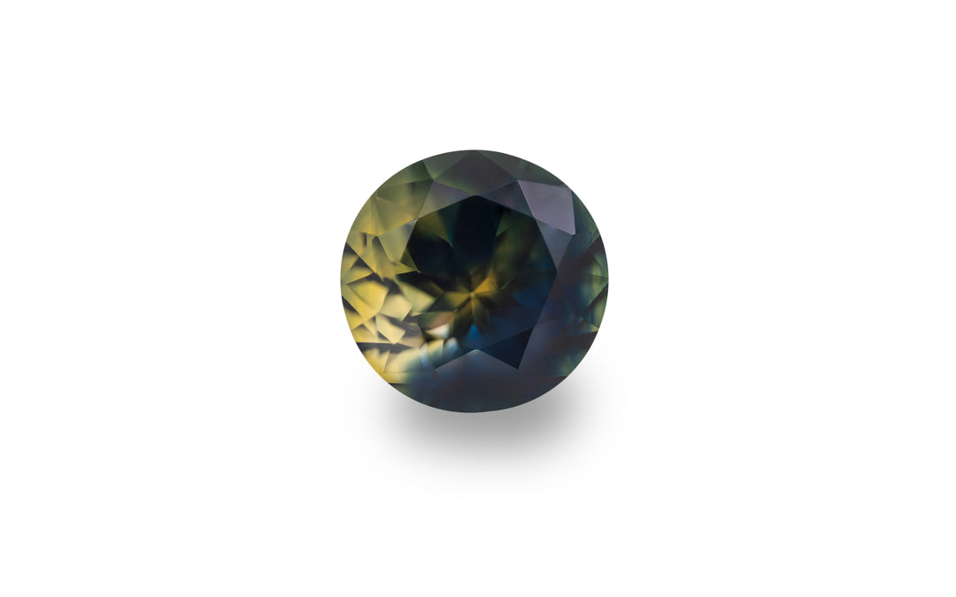 A round cut, royal blue, bright yellow and green Australian parti sapphire gemstone is displayed on a white background.