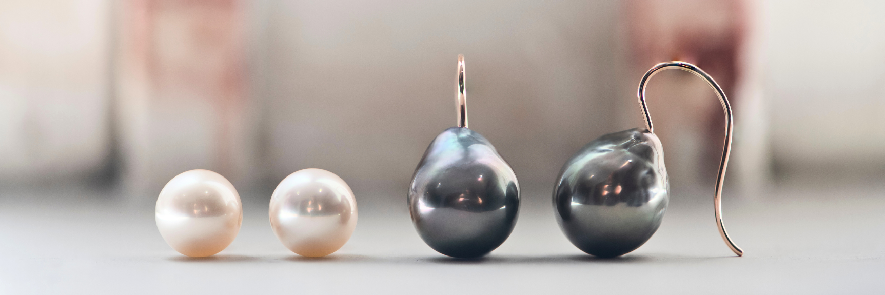 A pair of round white South Sea pearl stud earrings in 18K rose gold and a pair of Tahitian pearl baroque shepherd hook earrings in 18K rose gold are displayed on the edge of a windowsill.